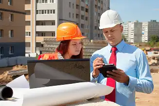 Two construction professionals on a site review plans on a laptop and a tablet near rolled blueprints and construction equipment at a building site with cranes in the distance
