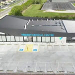 Aerial view of a modern medical clinic labeled 'Trident Medical Centre' with glass front, adjacent parking lot and accessible parking spaces in the foreground.