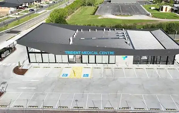 Aerial view of a modern medical clinic labeled 'Trident Medical Centre' with glass front, adjacent parking lot and accessible parking spaces in the foreground.