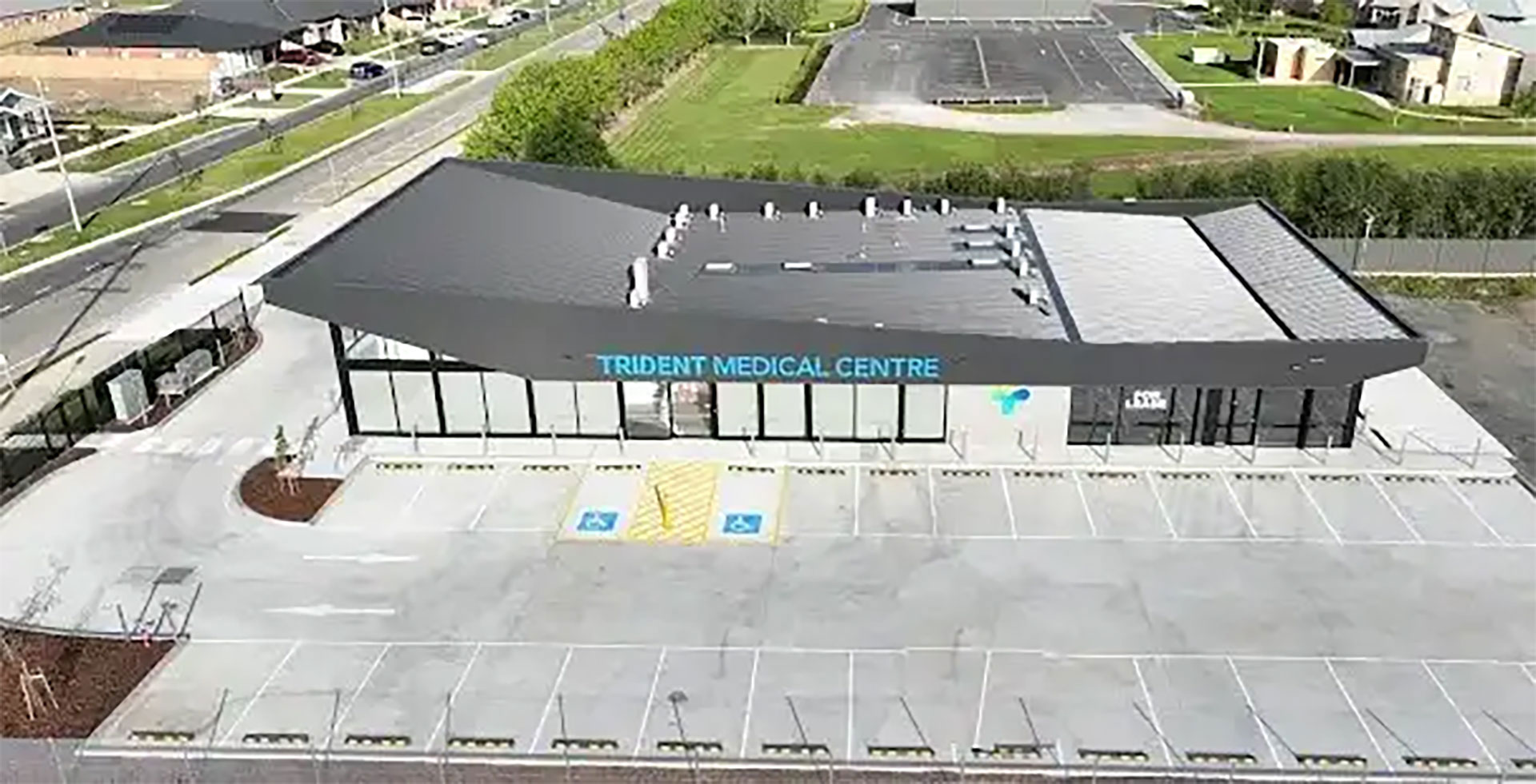 Aerial view of a modern medical clinic labeled 'Trident Medical Centre' with glass front, adjacent parking lot and accessible parking spaces in the foreground.