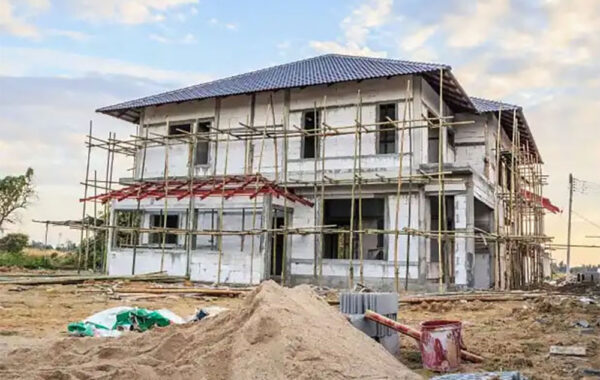 Two-story house under construction with bamboo scaffolding and a metal roof, construction debris in foreground.
