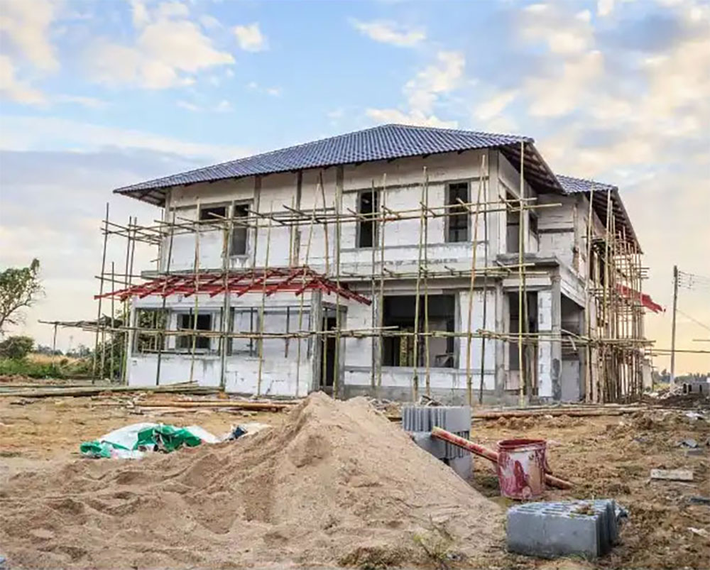 Two-story house under construction with bamboo scaffolding and a metal roof, construction debris in foreground.